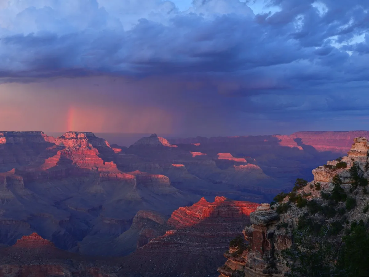 Late sunlight illuminating Grand Canyon formations beneath storm clouds and a rainbow near Yaki Point