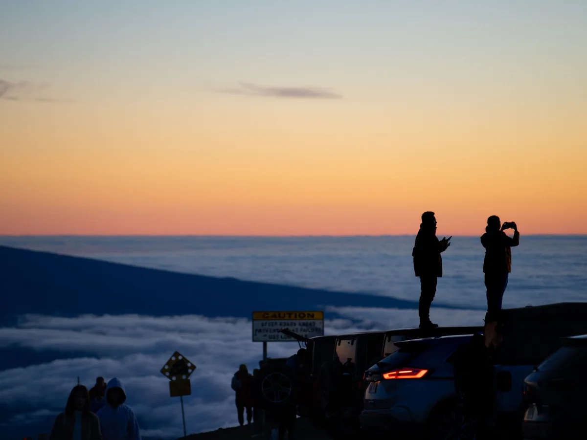 Visitors silhouetted at Haleakalā Summit above clouds during warm-to-blue twilight