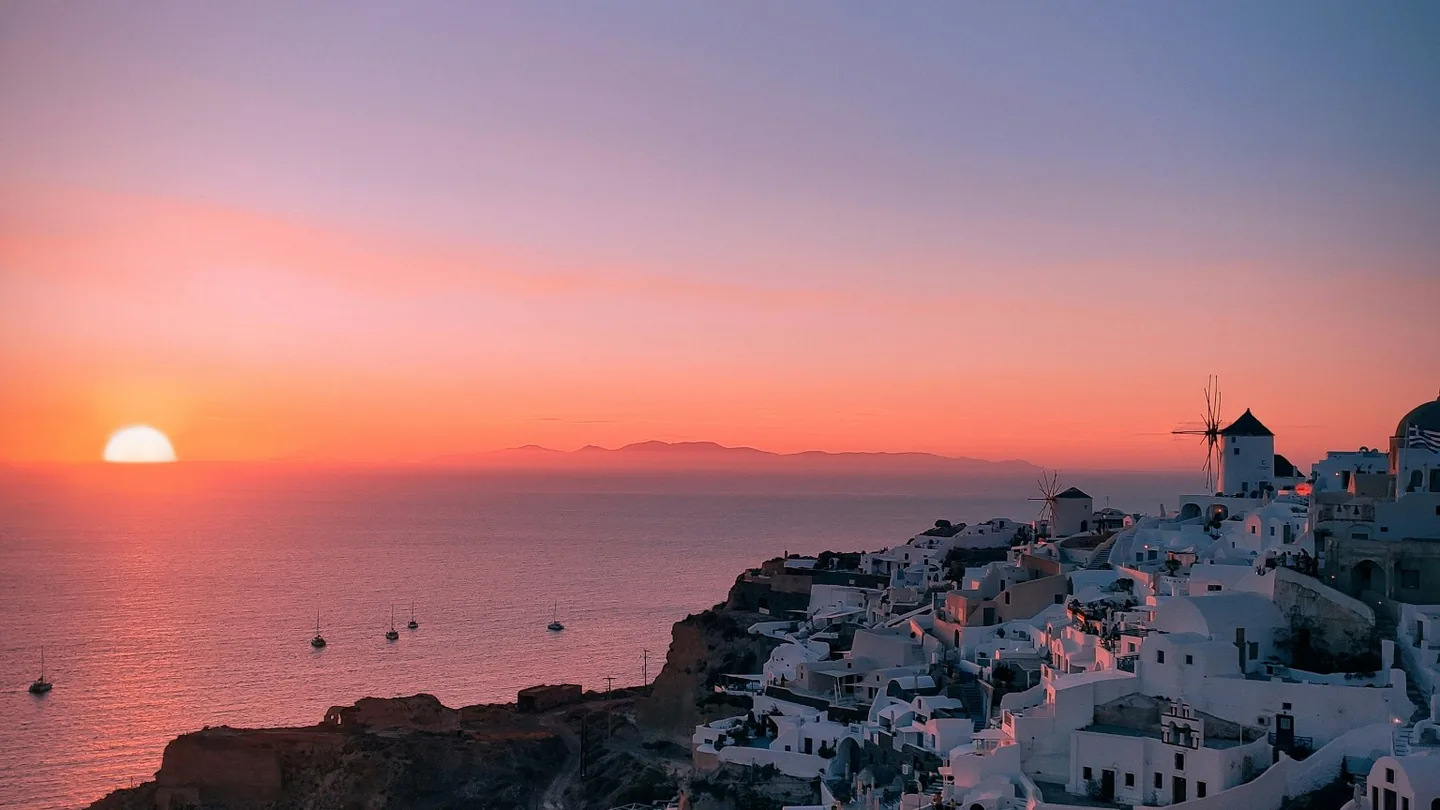 Sun setting over the Aegean beside Oia's whitewashed cliffside village and windmills in Santorini