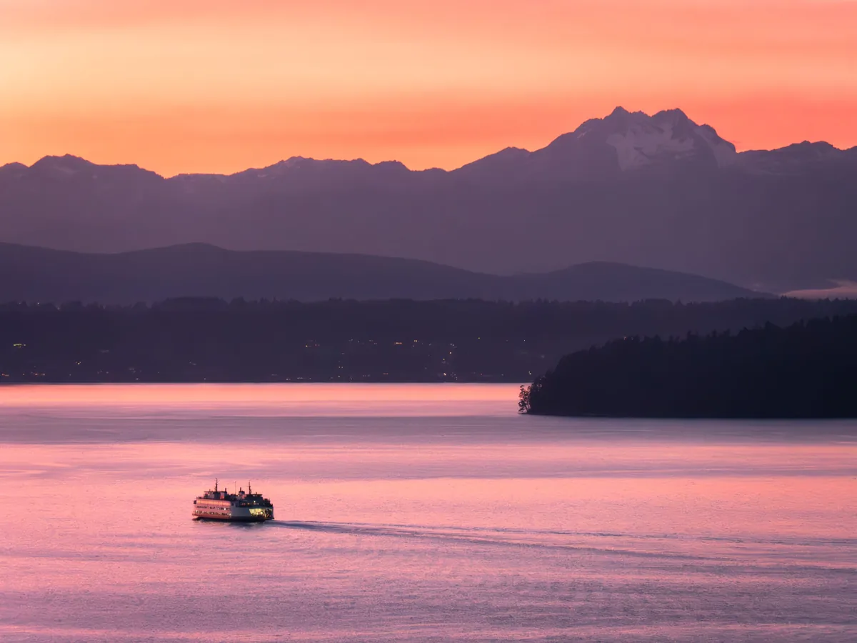 Ferry crossing Puget Sound beneath a pink sunset with the Olympic Mountains silhouetted in the distance
